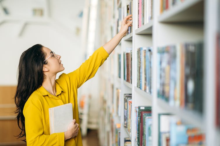 A woman wearing a yellow shirt reaches out for a book on a shelf.