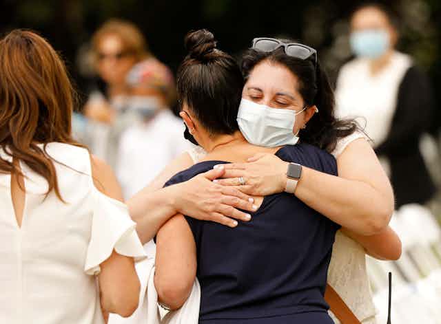 A brunette woman with a mask to protect against COVID-19 hugs a young woman in a black shirt.