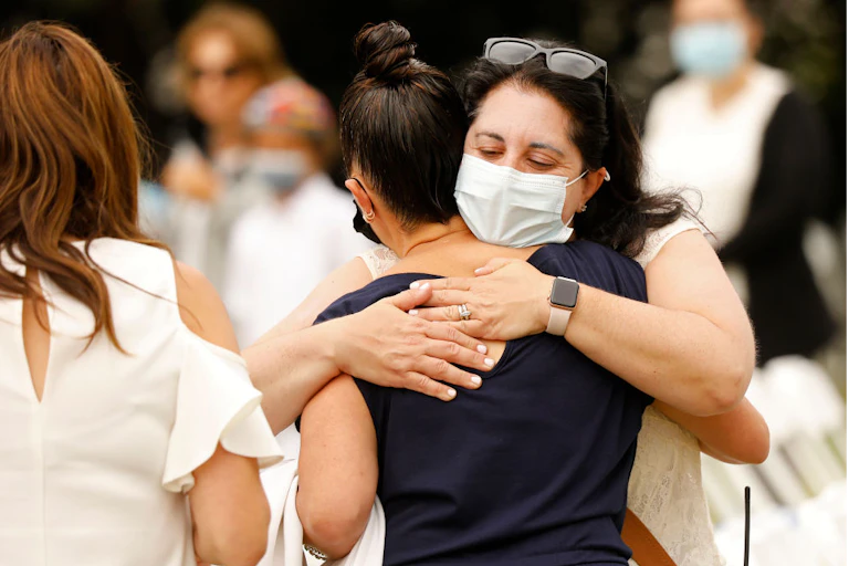 A brunette woman with a mask to protect against COVID-19 hugs a young woman in a black shirt.