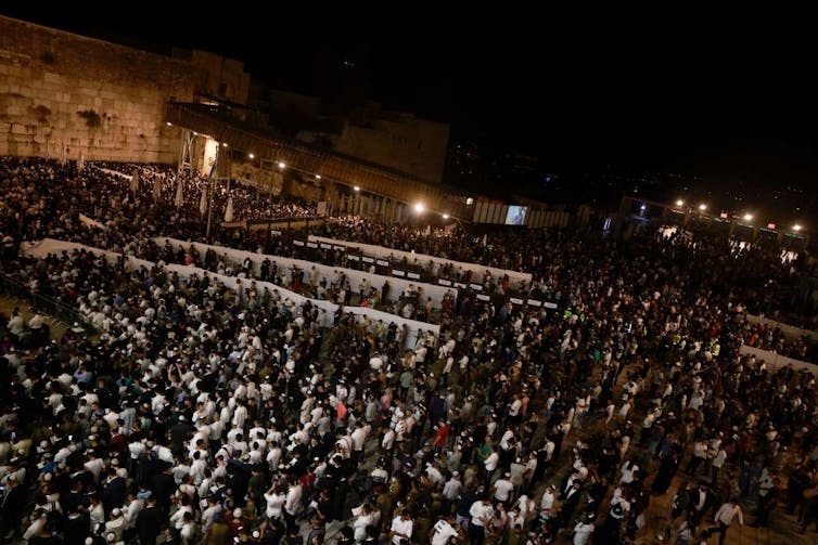 Many people dressed in black and white stand in a courtyard between ancient walls.