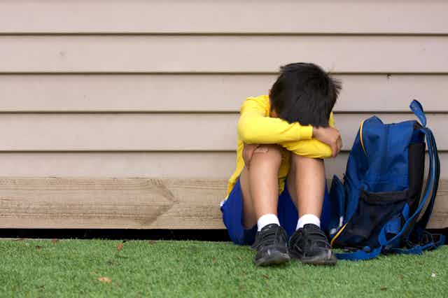 A young boy wearing a yellow shirt and blue shorts sits on grass with his back against the wall of a home with his head down as a blue backpack sits nearby.