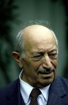 A color photograph of an older, balding man in a blue shirt and striped tie.