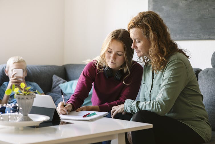 A mother helps her daughter do work as they sit on the couch and work on a notepad that lies on a nearby table.