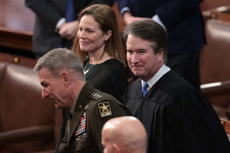 A white woman with brown hair and a black dress stands next to a man in military uniform, who stands next to another white man with a black robe.