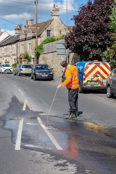 A man in a reflective orange jacket locating a water leak on a town road.