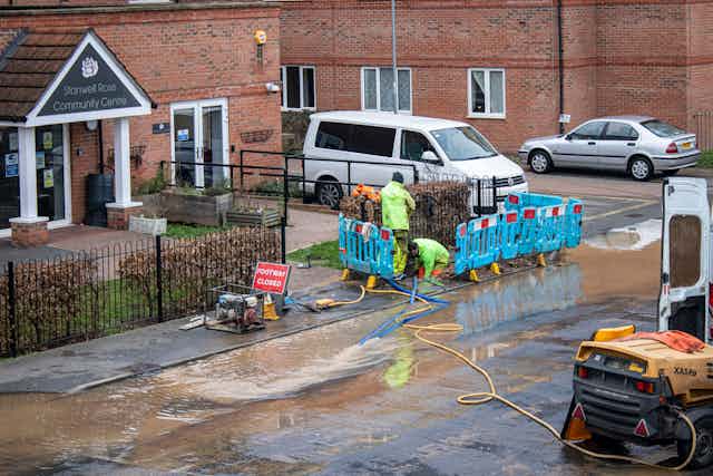 A large puddle covering an urban roadside, with two water engineers crouched down fixing the leak.