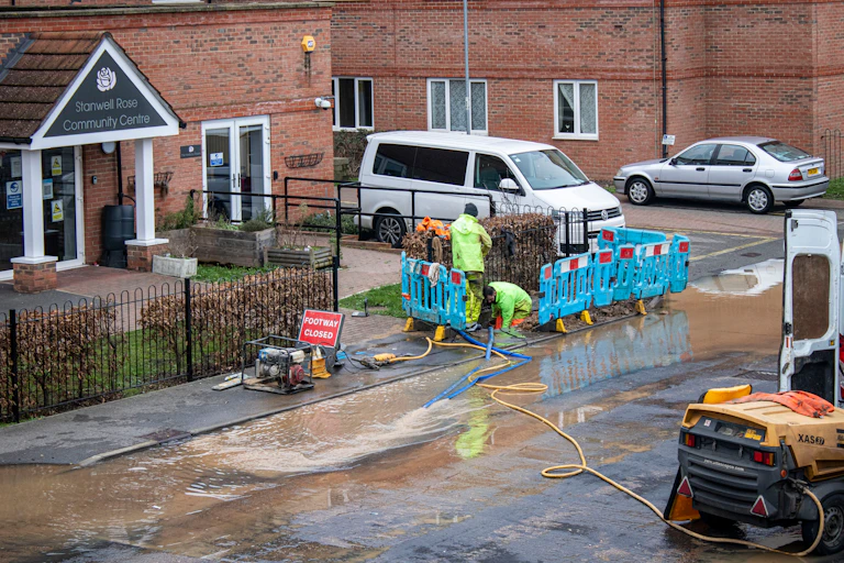 A large puddle covering an urban roadside, with two water engineers crouched down fixing the leak.