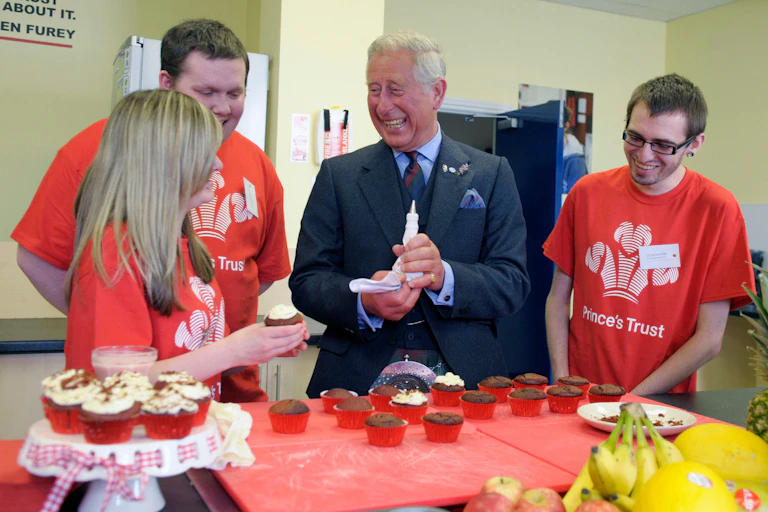 Prince Charles laughs while decorating cupcakes with there young people in red Prince's Trust t-shirts.