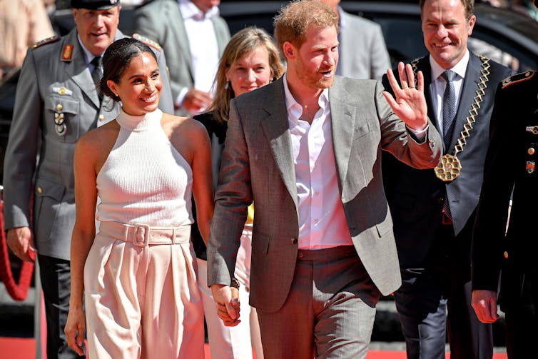 Prince Harry and Meghan Markle, smiling and holding hands, arrive at an Invictus Games Foundation event, Harry is waving to supporters