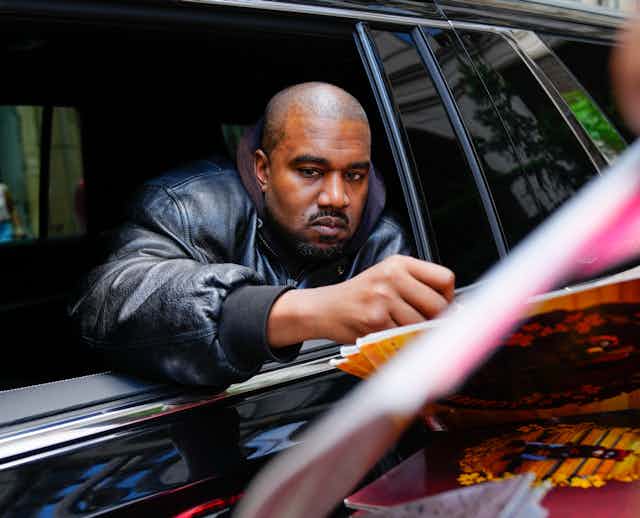 Kanye West signs an autograph while his arm is extended over a rolled-down window from inside a black vehicle.