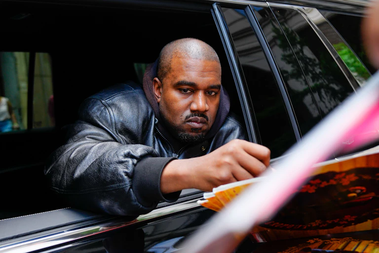 Kanye West signs an autograph while his arm is extended over a rolled-down window from inside a black vehicle.