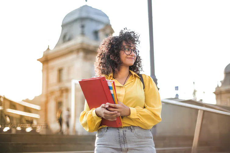 A woman seen in a yellow shirt on a campus holding books and smiling.