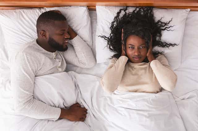 A young couple in bed. One has their hands over their ears because their partner is snoring.