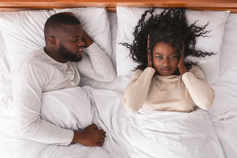 A young couple in bed. One has their hands over their ears because their partner is snoring.
