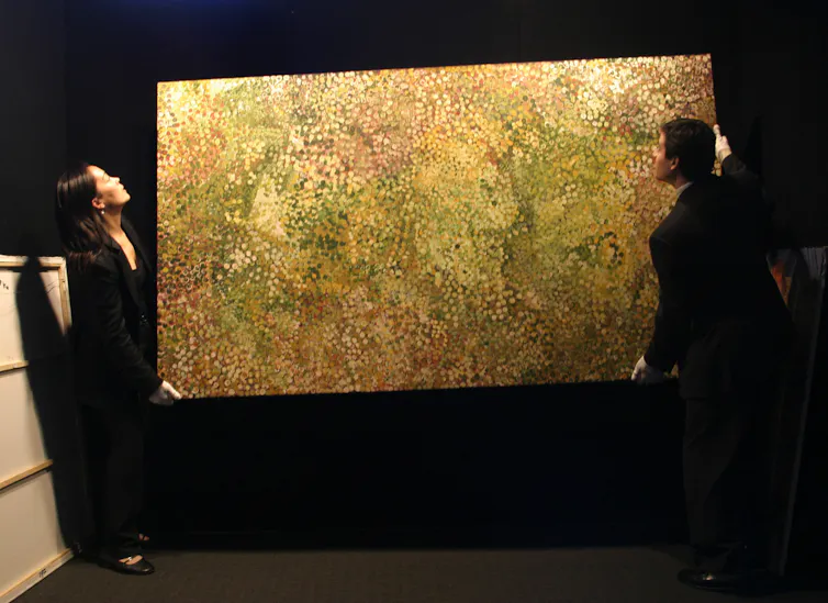 two black-clad people moving a large painting