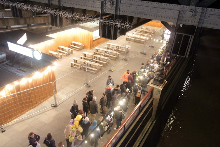 A queue of people under a bridge at nighttime with bright lights.