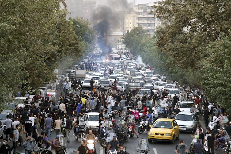 A large crowd and cars are seen on a tree-lined city street, smoke billowing in places.
