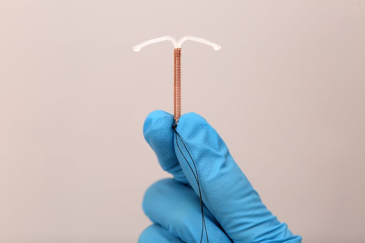 A clinician wearing blue surgical gloves holds a copper IUD between their fingers.