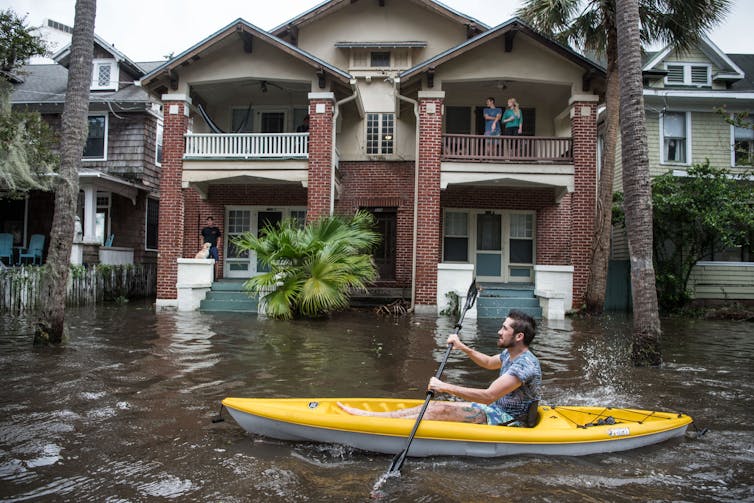 A man kayaks through a flooded street, past people standing on porches with water up the stairs of the homes. The yards aren't visible.