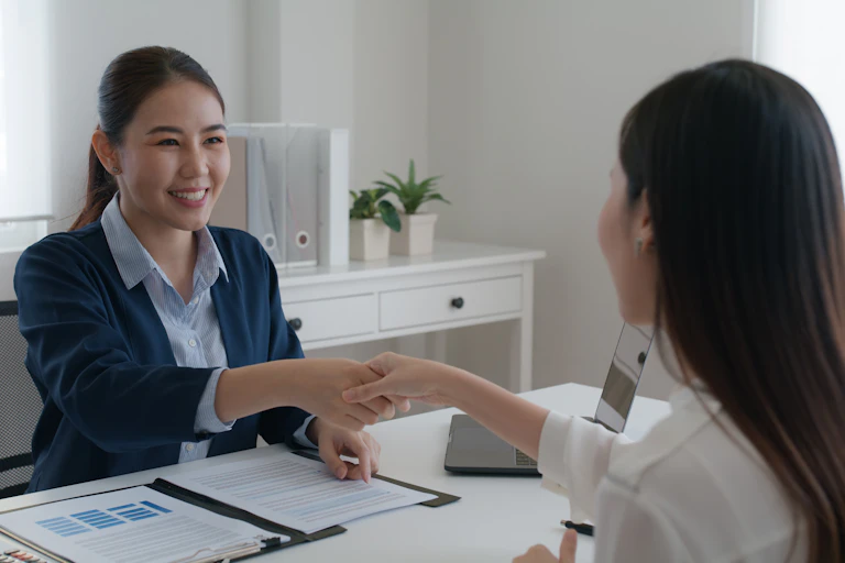 Two Chinese women smile and shake hands across a desk with financial charts and documents on the desk