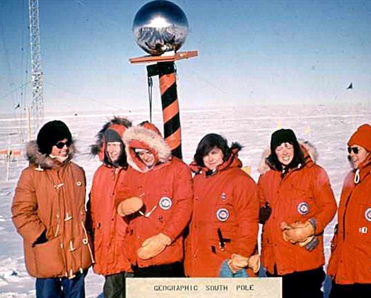 Six women wearing heavy parkas stand in front of a large striped pole with a mirrored ball on top.