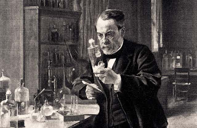 Standing next to a table covered by test tubes and beakers, Louis Pasteur holds a bottle partially filled with an unidentified liquid.