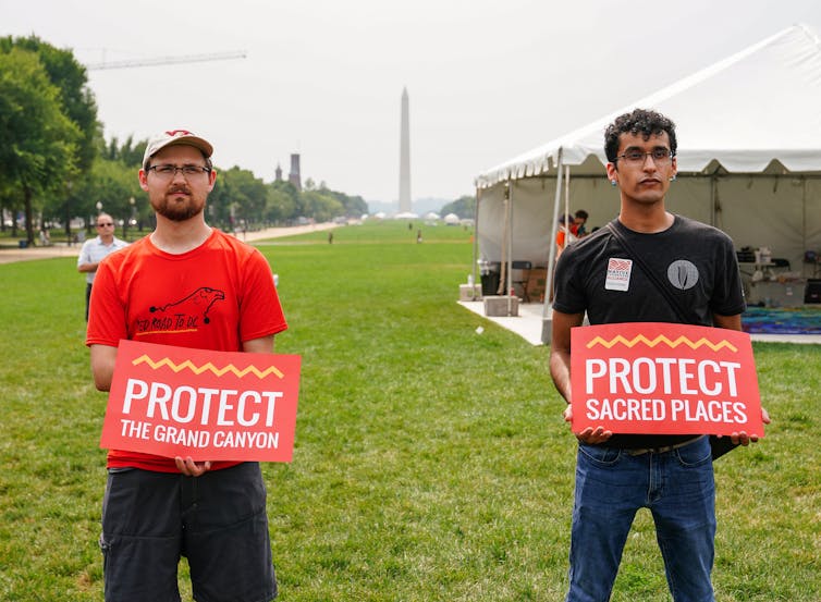 Two men stand with signs reading, 'Protect sacred places' on the National Mall, with the Washington Monumemt visible in the background.