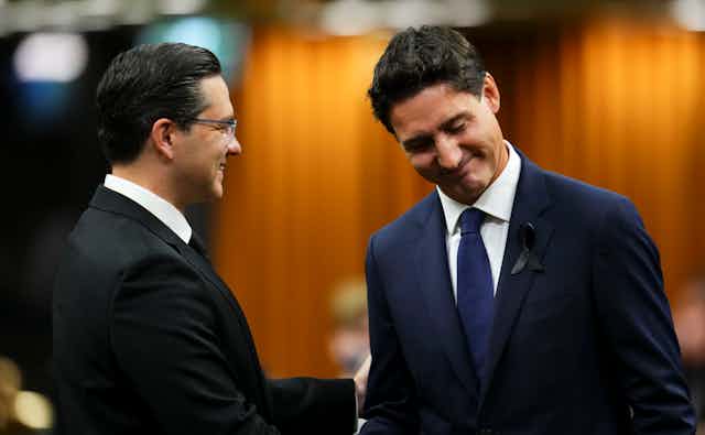 Two men in dark suits, one in glasses, shake hands. Both are smiling, one looking down.