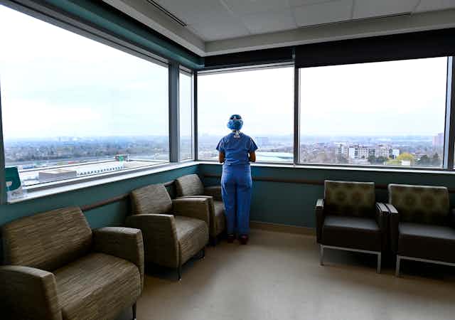 A person in blue scrubs seen from behind, looking out a large window