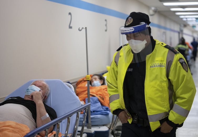 A paramedic in a face shield wearing a neon yellow jacket walks past patients on gurneys in a hospital corridor