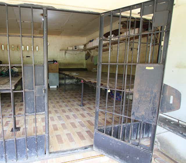 A prison door with bars is left open to reveal a small cell that includes several wooden tables and thin mattresses.