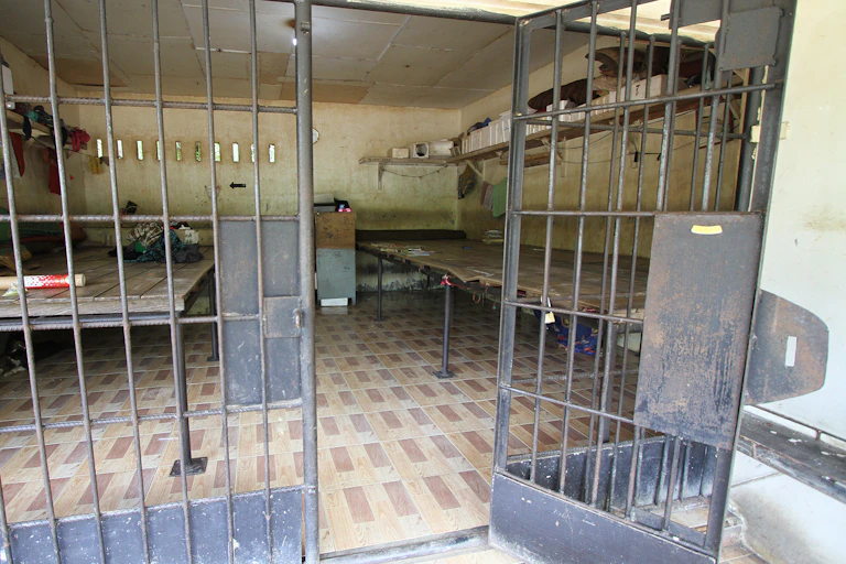 A prison door with bars is left open to reveal a small cell that includes several wooden tables and thin mattresses.