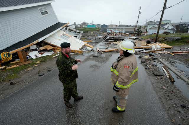 a firefighter and army officer stand in the middle of a street surrounded by debris from a destroyed home