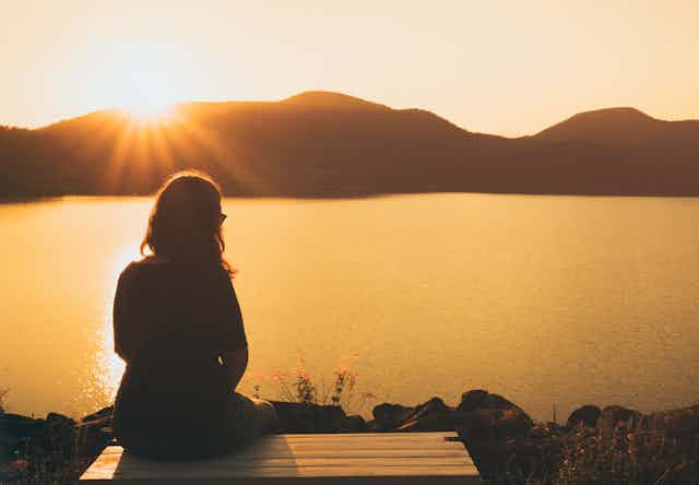 Silhouette of a person sitting near water, with the sun rising over some hills