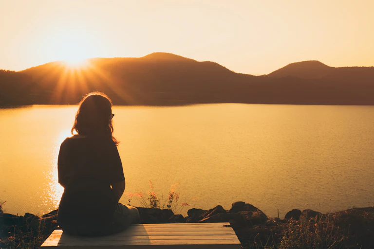 Silhouette of a person sitting near water, with the sun rising over some hills