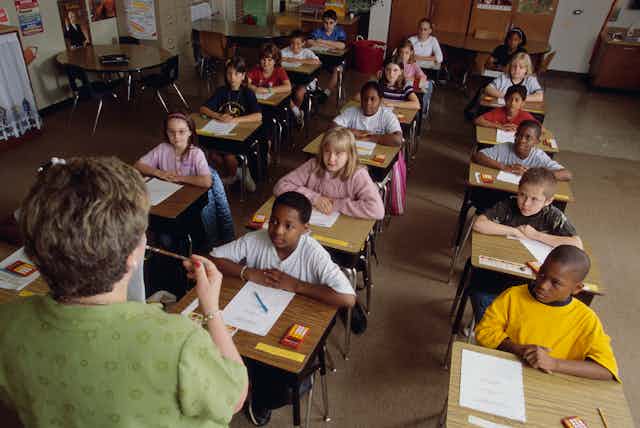 A white female schoolteacher stands in front of an elementary class of students from different racial and ethnic backgrounds.