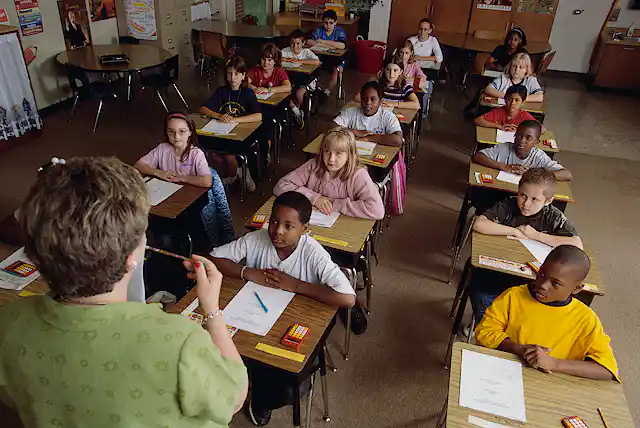 A white female schoolteacher stands in front of an elementary class of students from different racial and ethnic backgrounds.