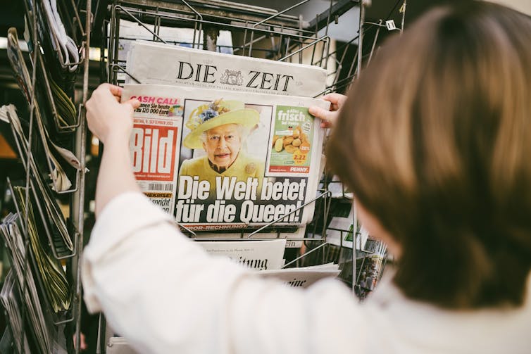 A woman takes a German newspaper off of a stand, the front page has a photo of the Queen and a headline reading Die Welt betet fur die Queen.