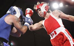 Two men, one in blue boxing gear and the other in red, fight in a boxing ring.
