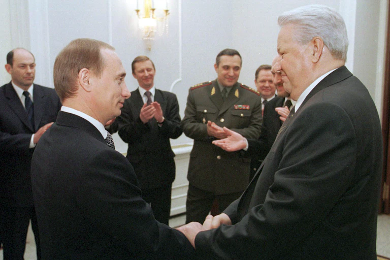 Vladimir Putin shakes hands with Boris Yeltsin as ministers applaud.