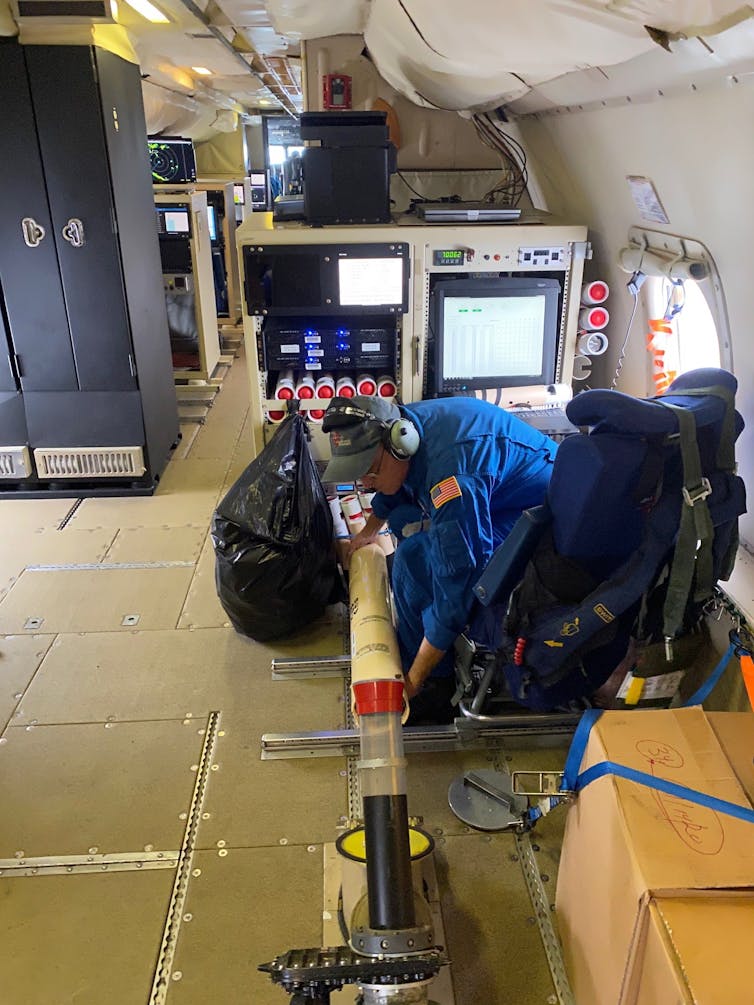 A scientist in a flight suit puts a device into a tube in the bottom of the plane to drop it.