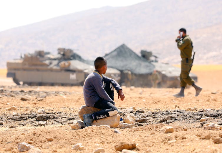 A boy sitting in front of two tanks and a soldier