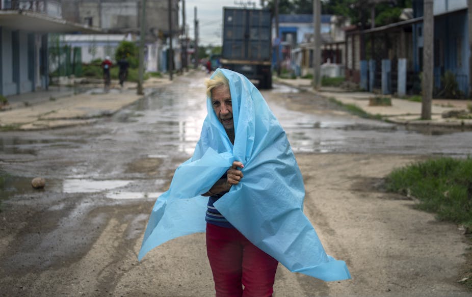 An older woman with plastic sheeting wrapped around herself walks through a street during a storm.