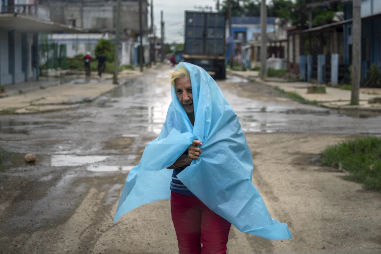 An older woman with plastic sheeting wrapped around herself walks through a street during a storm.
