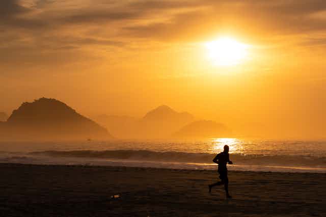 A man jogs on a beach on a hazy day with the sun over the water.