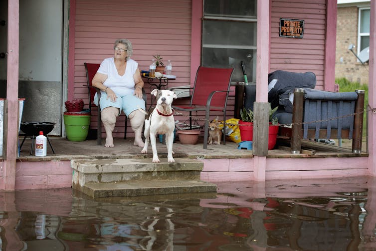 A woman sits on a porch with a dog as floodwater covers the stairs. A sign reading'private property: no tresspassing' is on the house.