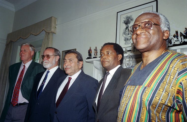 A row of men wearing suits and one wearing a traditional African shirt stand in a row.