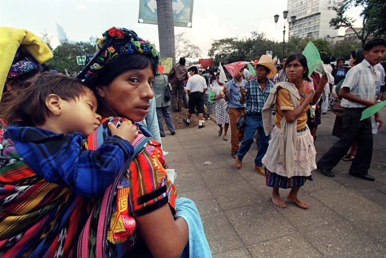 A woman wearing bright-colored clothing holds a sleeping child on her back while she watches a parade of people walking down a street.