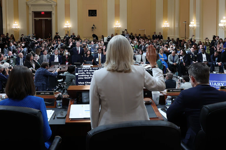 The back of a blond woman is seen raising her hand, facing a monitor, as she appears to be facing a man in a suit with his hand raised. They are in a crowded room with people seated in formal clothing.