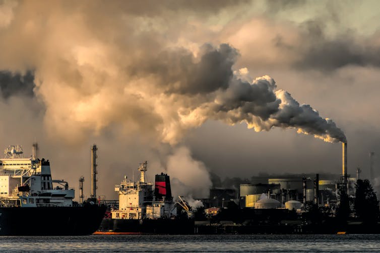 Tankers in the foreground of an industrial landscape. Billowing emissions from smokestacks rise in the air.